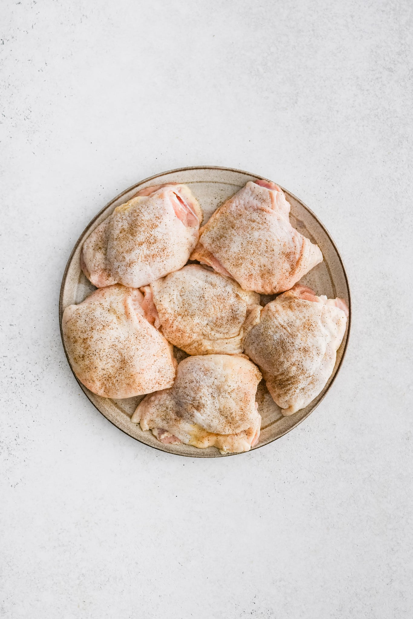 Six raw bone-in skin-on chicken thighs seasoned with salt and pepper on a plate.