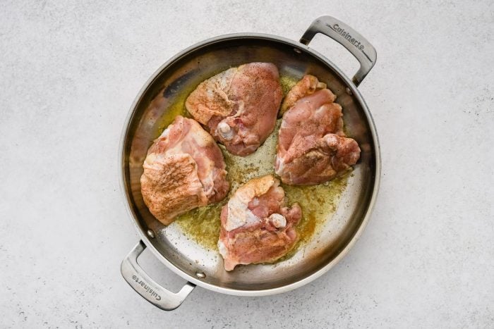 Four bone-in skin-on chicken thighs browning in a large pan skin-side-down.