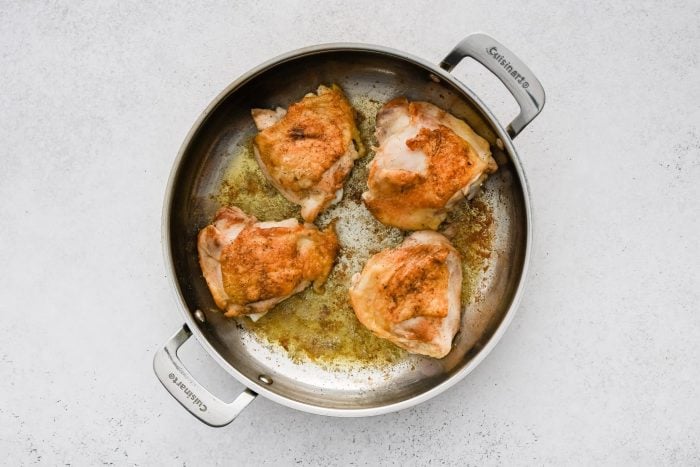 Four bone-in skin-on chicken thighs browning in a large pan skin-side-up.