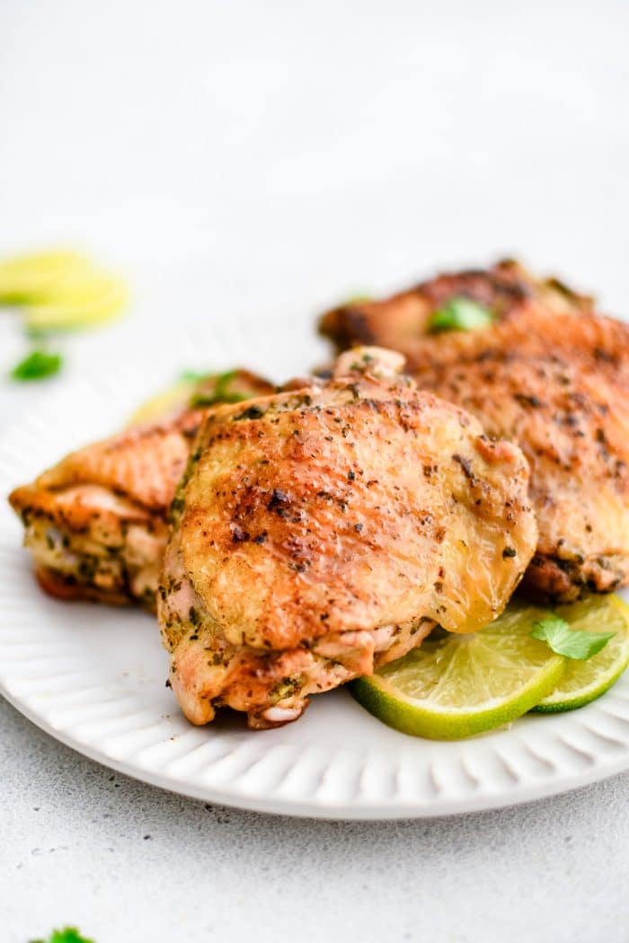 Large white serving plate topped with four juicy cooked chicken thighs marinated in cilantro lime marinade and served with  fresh lime slices.