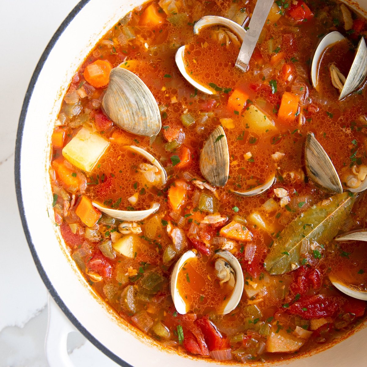 Overhead image of a large pot of Manhattan clam chowder filled with carrots, celery, onion, potatoes, little neck clams, all simmering in a tomato and clam broth.