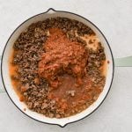 Large skillet filled with cooked ground beef and softened onion with homemade tomato sauce made from blended garlic cloves, jalapeno, tomatoes, whole pieces of onion, oregano, beef bouillon, salt, pepper, ground cumin, and two cups of water.