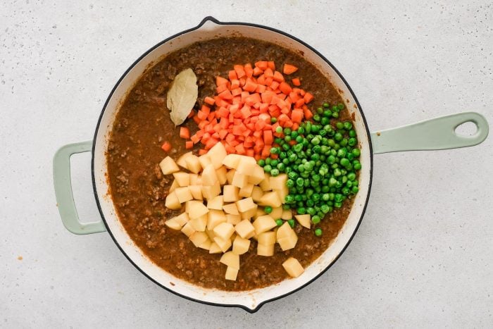 Diced potatoes, carrots, and peas added to a large skillet filled with simmering ground beef in homemade tomato sauce.