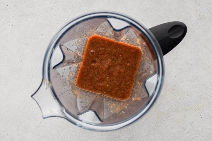 Blender filled with pureed garlic cloves, jalapeno, tomatoes, whole pieces of onion, oregano, beef bouillon, salt, pepper, ground cumin, and two cups of water.