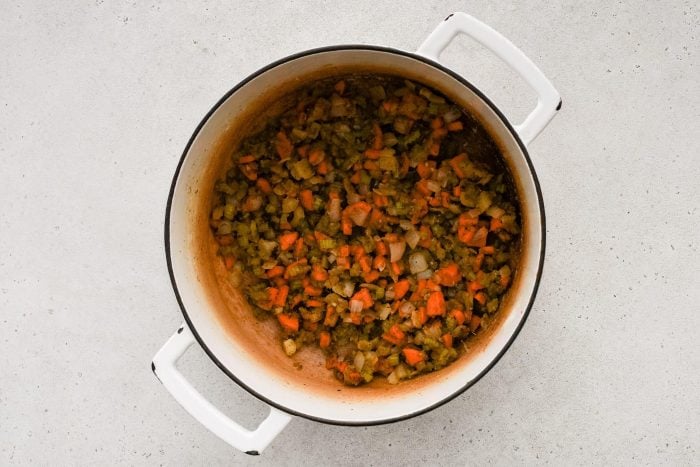 Large white dutch oven over medium heat filled with diced pieces of onion, carrot, and celery coated in flour, garlic, thyme, pepper, and salt.