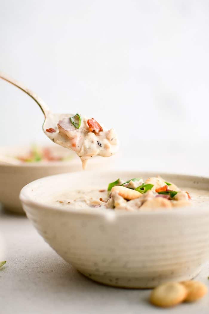 Spoon and bowl filled with homemade New England Clam Chowder garnished with oyster crackers, green onion, and bacon bits.