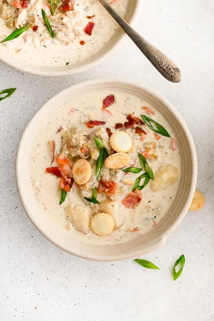 White soup bowl filled with homemade New England Clam Chowder garnished with oyster crackers, green onion, and bacon bits.