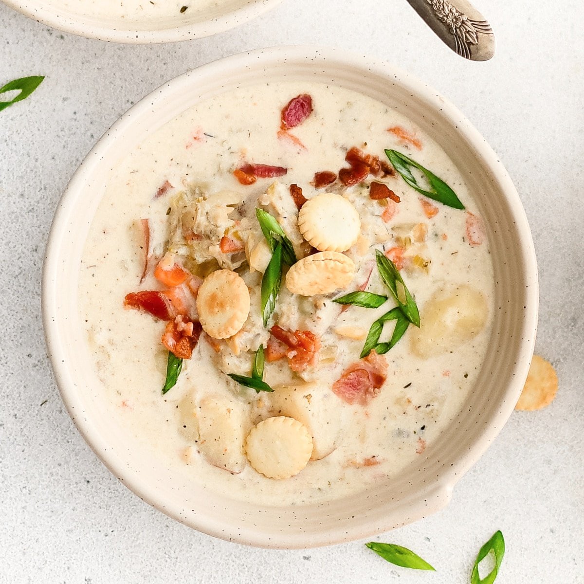 White soup bowl filled with homemade New England Clam Chowder garnished with oyster crackers, green onion, and bacon bits.