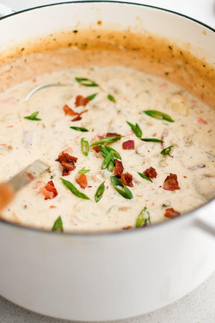 Large white Dutch oven filled with creamy New England clam chowder garnished with sliced green onions and bacon bits.