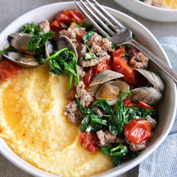 Large shallow serving bowl filled with creamy polenta and topped with cooked ground Italian sausage with sauteed spinach and cherry tomatoes and cooked little neck clams.