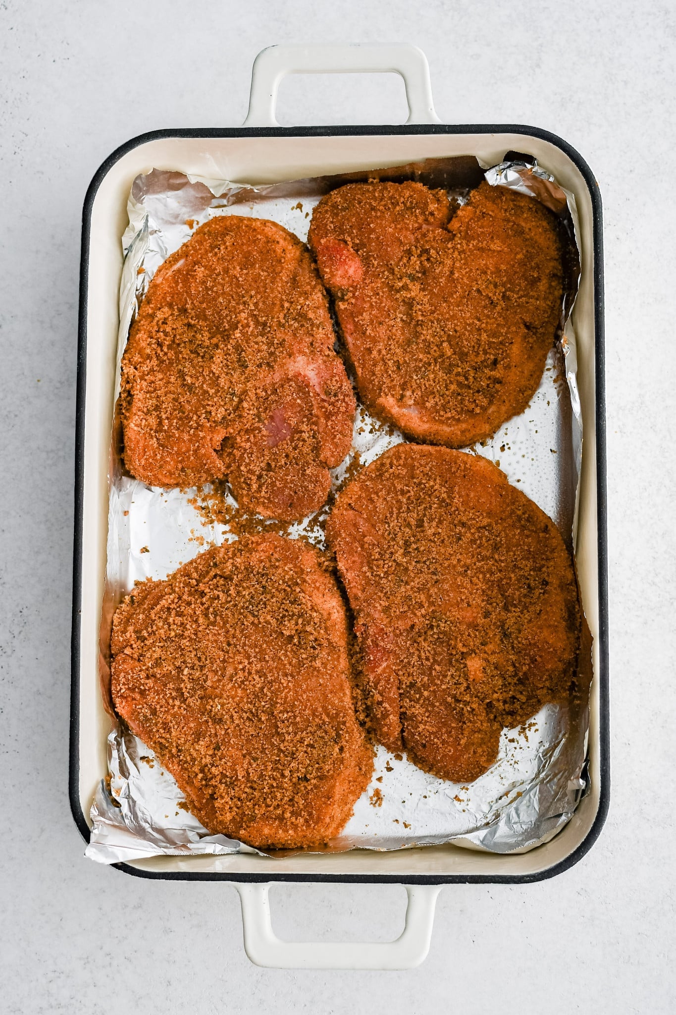 Four raw pork chops seasoning with homemade shake and bake in a large baking dish lined with foil.