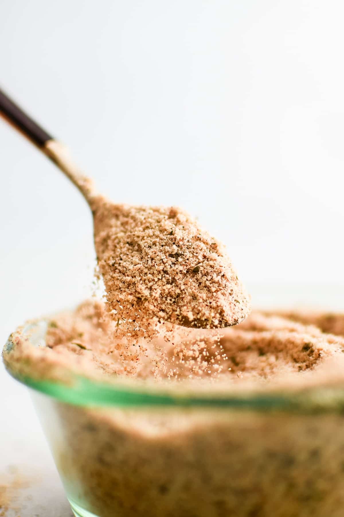 Spoon filled with homemade Shake and Bake seasoning spilling from the edges into a glass jar filled with the rest of the seasoning.