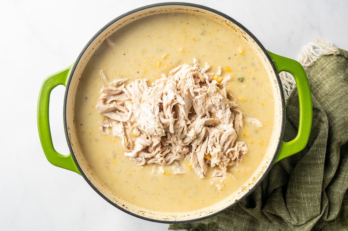A generous pile of shredded chicken added to the center of creamy corn chowder with visible pieces of corn and vegetables, ready to be stirred back into the soup.