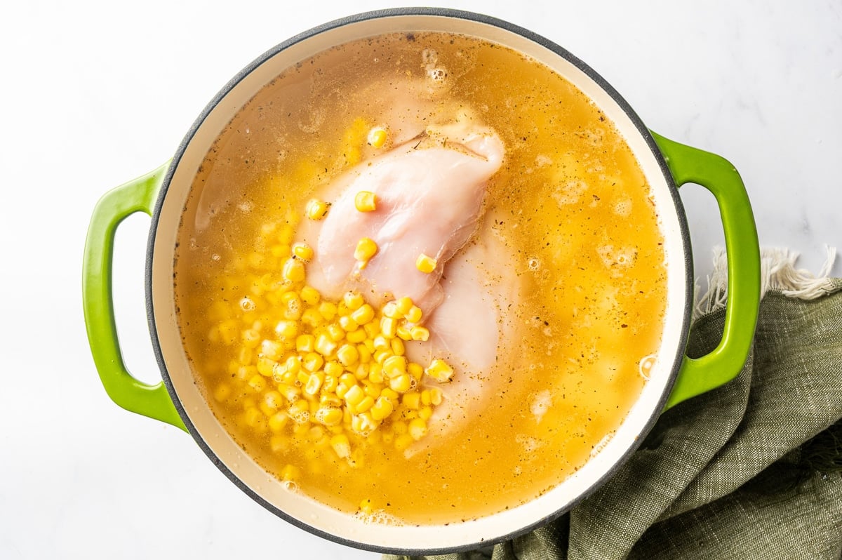 Boneless skinless chicken breasts and bright yellow sweet corn submerged in seasoned broth in a green Dutch oven as the chicken corn chowder simmers.