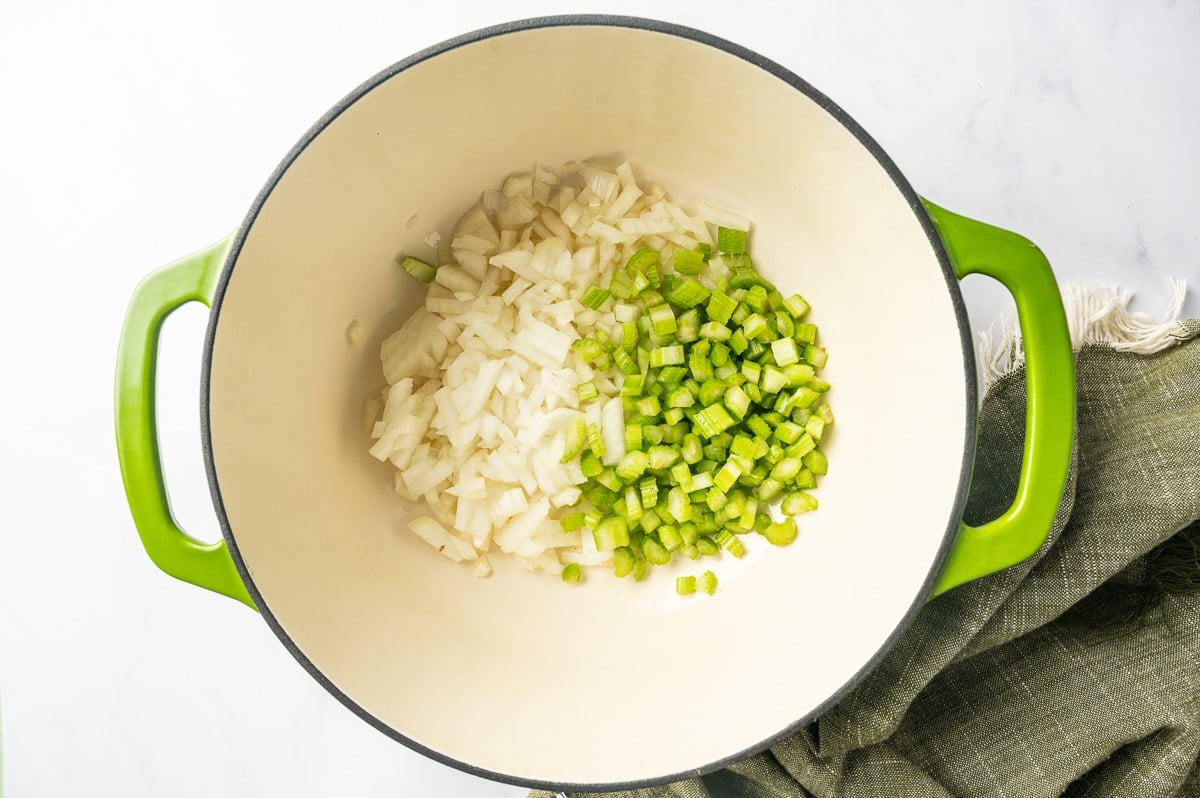 Chopped white onion and celery cooking together in a green enameled Dutch oven as the vegetable base for chicken corn chowder.