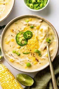 Creamy chicken corn chowder served in a wide bowl with pieces of shredded chicken and sweet corn throughout, topped with cheddar cheese, sliced jalapeno, green onions, and black pepper, with a spoon resting in the bowl.
