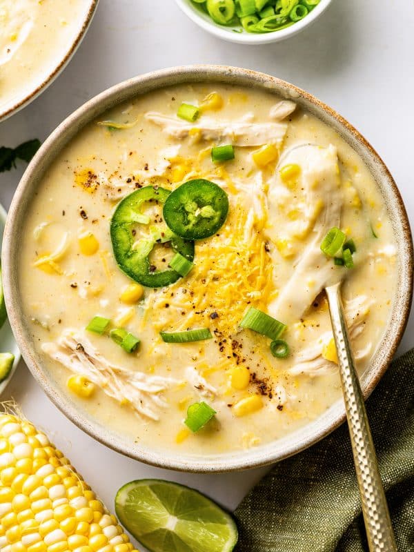 Creamy chicken corn chowder served in a wide bowl with pieces of shredded chicken and sweet corn throughout, topped with cheddar cheese, sliced jalapeno, green onions, and black pepper, with a spoon resting in the bowl.