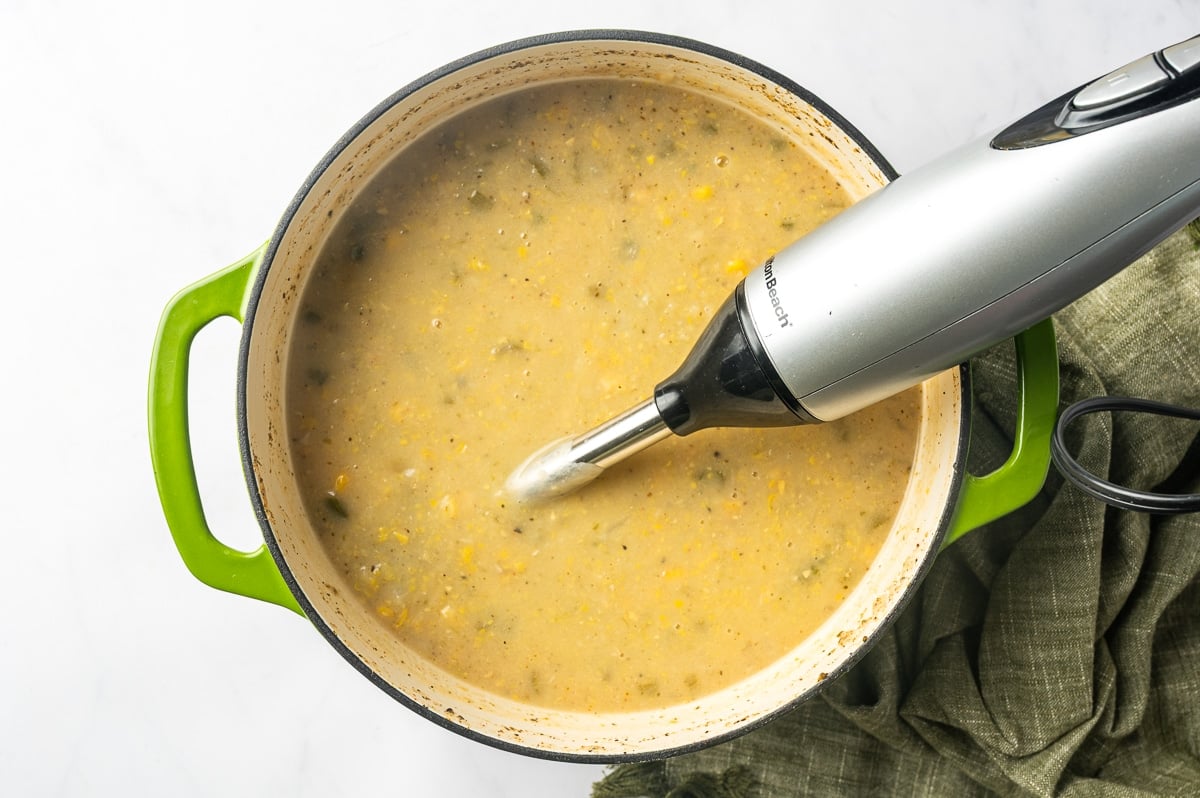 An immersion blender partially blending the corn, potatoes, vegetables, and broth in a Dutch oven to create a thicker, creamier chicken corn chowder base.