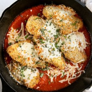 Large cast iron skillet filled with homemade marinara sauce and overlapping crispy coated baked chicken cutlets topped with melted cheese and fresh chopped basil.