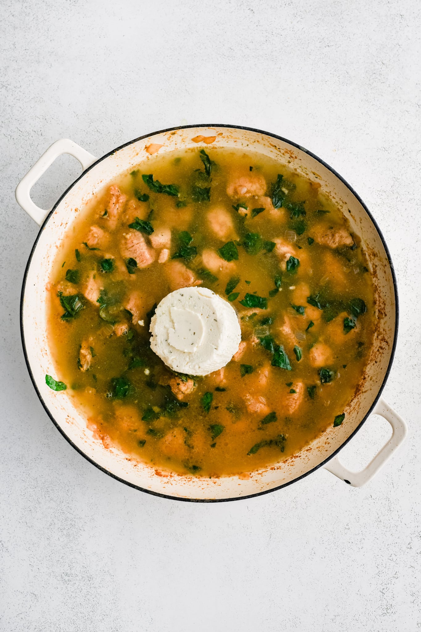 One wheel of Boursin cheese and chicken broth added to a large pan filled with cooked cubes of seasoned chicken breast, softened diced onion, and wilted spinach.