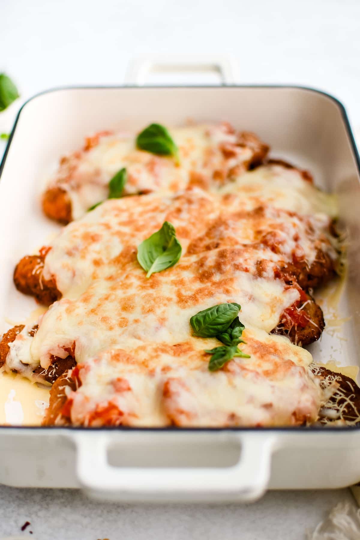 Large baking dish with breaded fried chicken cutlets topped with homemade tomato sauce and melted cheese and fresh basil leaves.