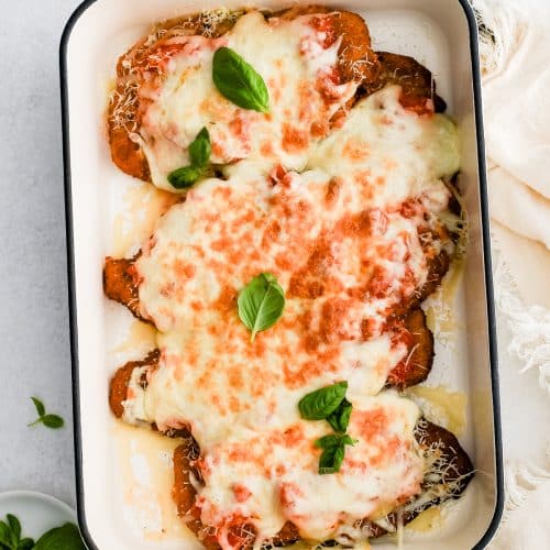 Large baking dish with breaded fried chicken cutlets topped with homemade tomato sauce and melted cheese and fresh basil leaves.