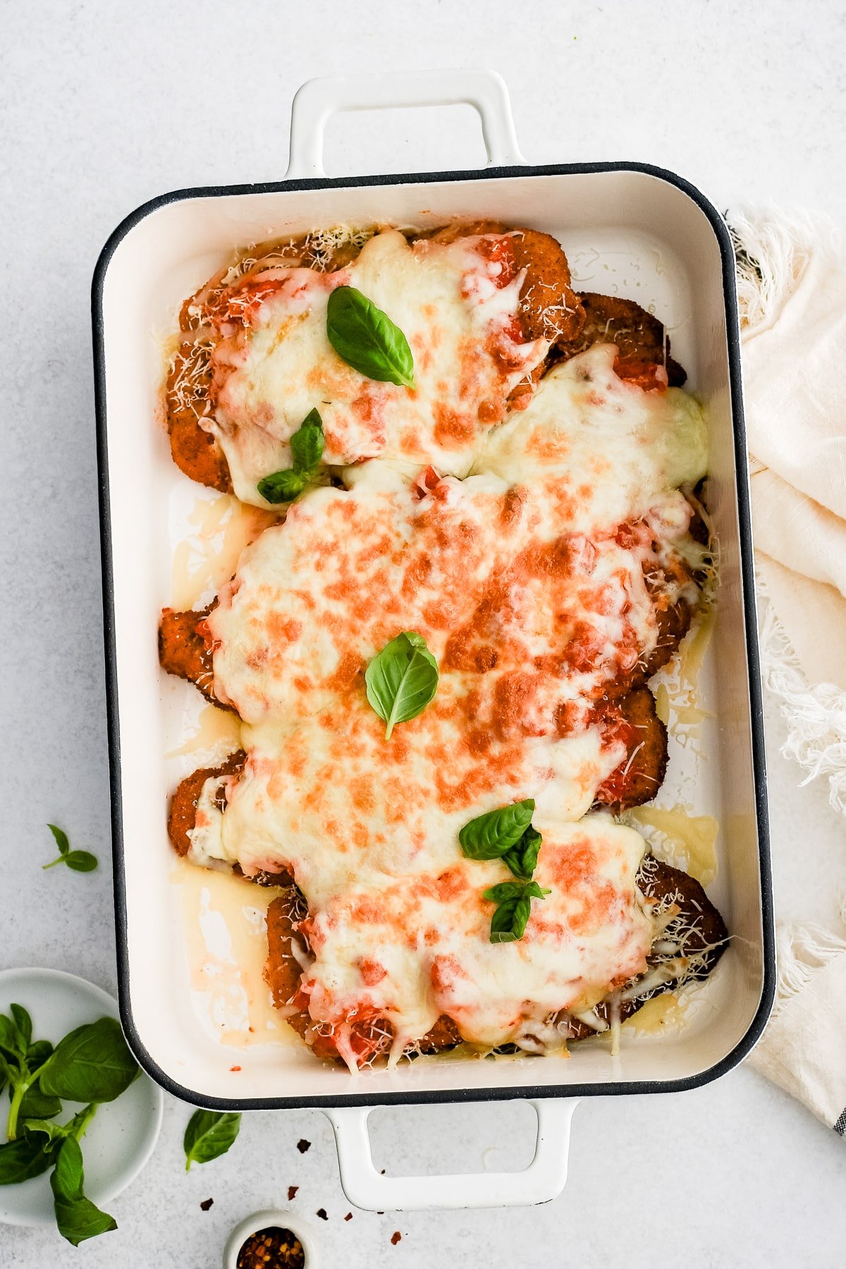 Large baking dish with breaded fried chicken cutlets topped with homemade tomato sauce and melted cheese and fresh basil leaves.