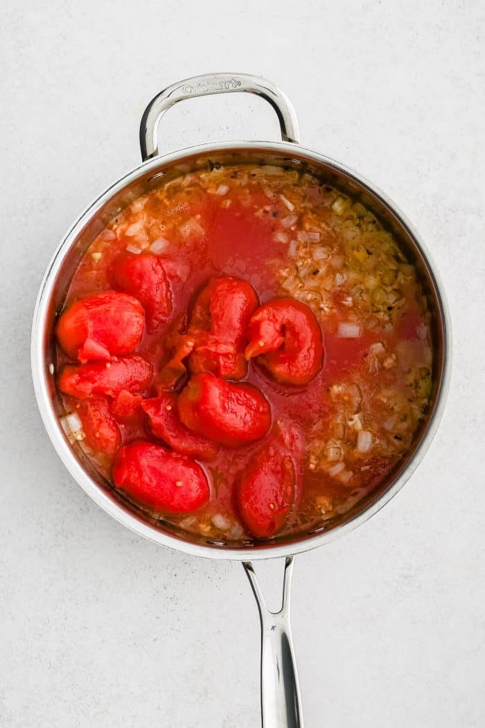 Can of whole tomatoes added to a pot filled with softened onion, garlic, salt, and pepper.