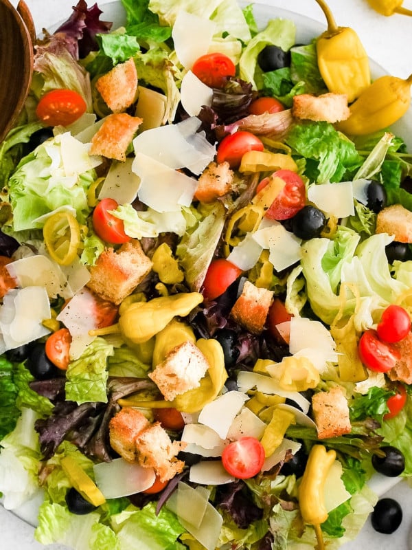 Round serving plate with crisp Romaine lettuce, iceberg, radicchio, halved cherry tomatoes, black olives, sliced pepperoncini, shaved parmesan, and croutons.