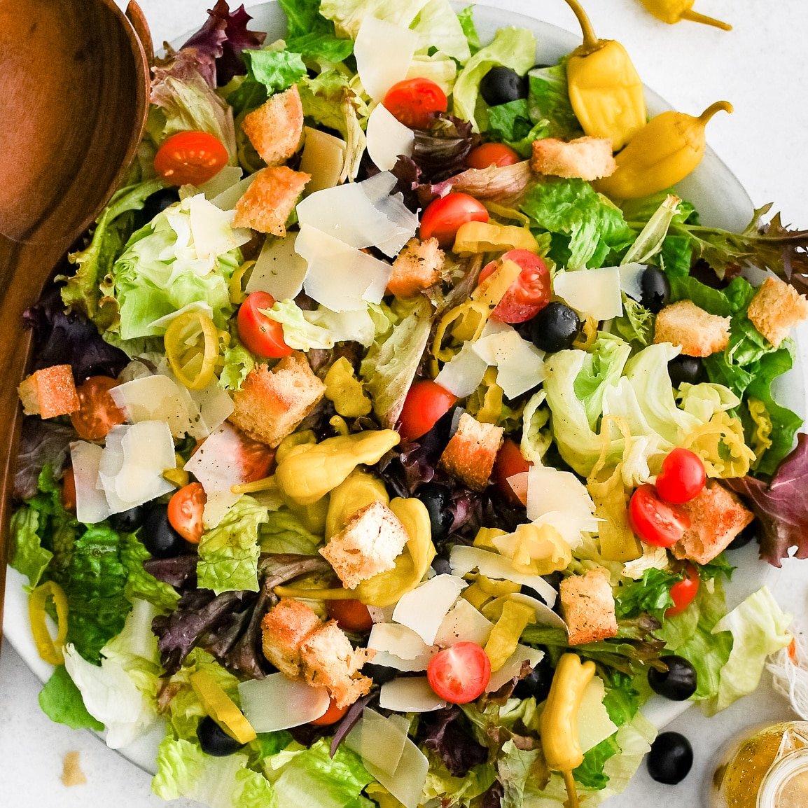 Round serving plate with crisp Romaine lettuce, iceberg, radicchio, halved cherry tomatoes, black olives, sliced pepperoncini, shaved parmesan, and croutons.