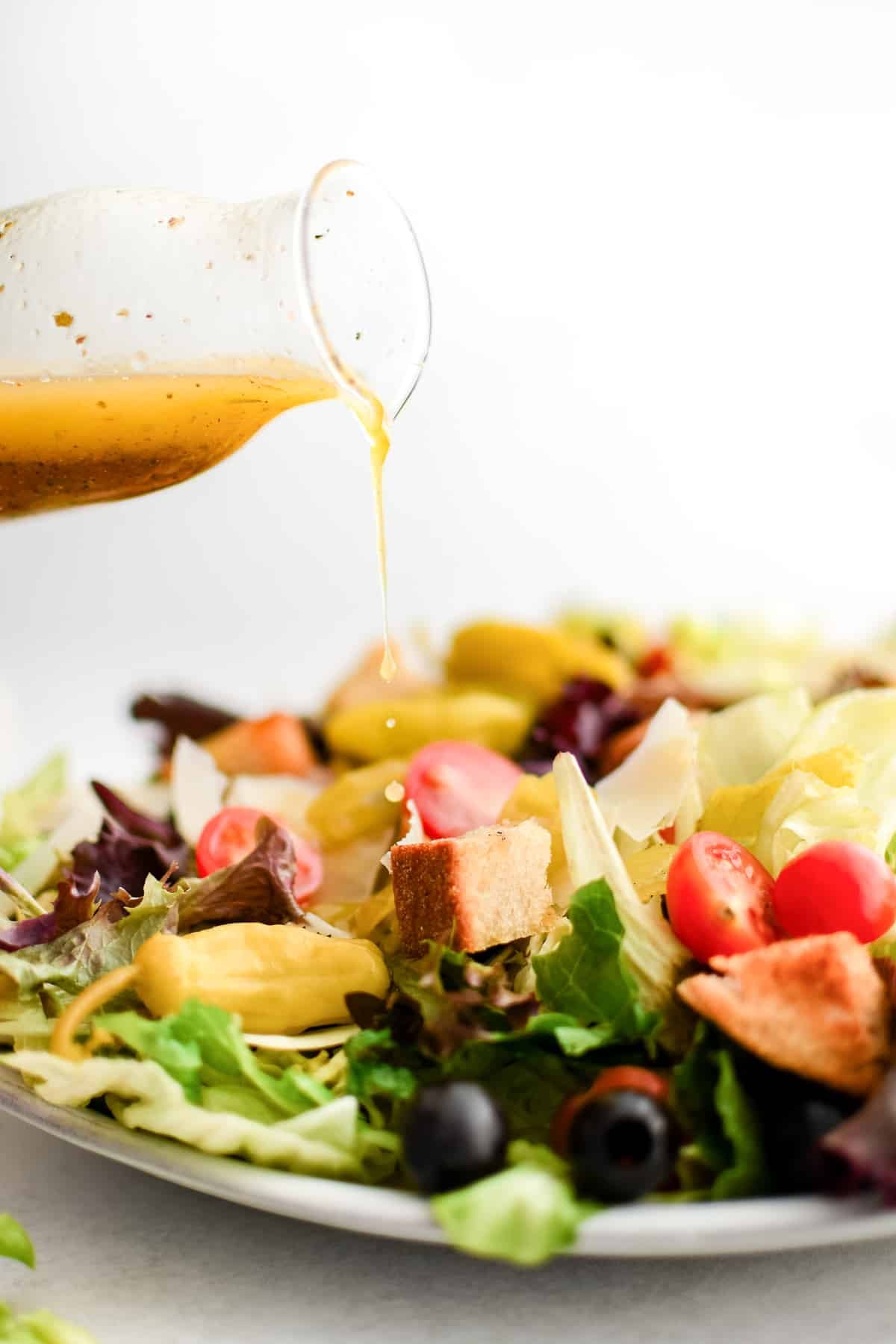 Drizzling homemade Italian dressing over a large Italian salad consisting of crisp Romaine lettuce, iceberg, radicchio, halved cherry tomatoes, black olives, sliced pepperoncini, shaved parmesan, and croutons.