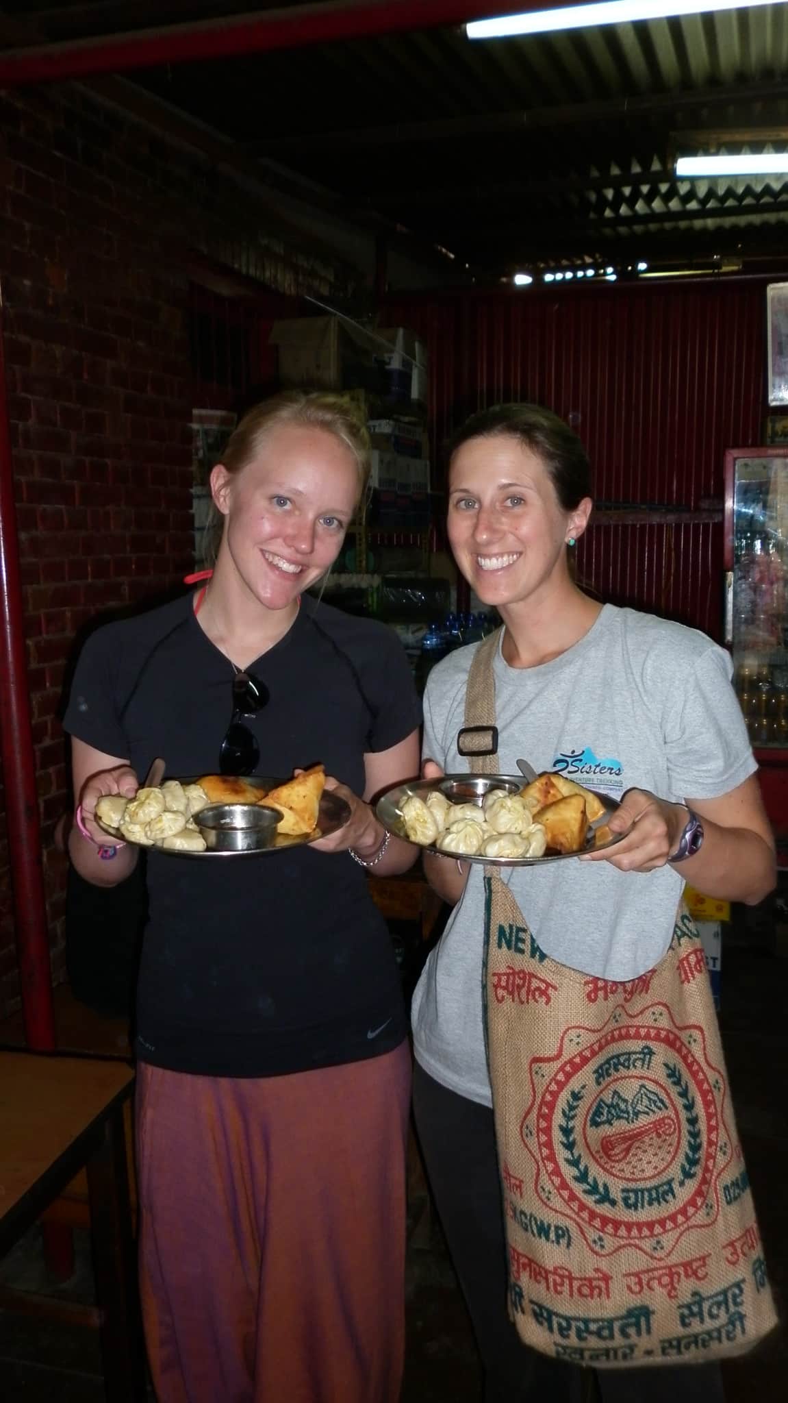 Jessica Randhawa and a friend at a cooking school in Kathmandu, Nepal, holding plates of freshly made momo dumplings and samosas after class.