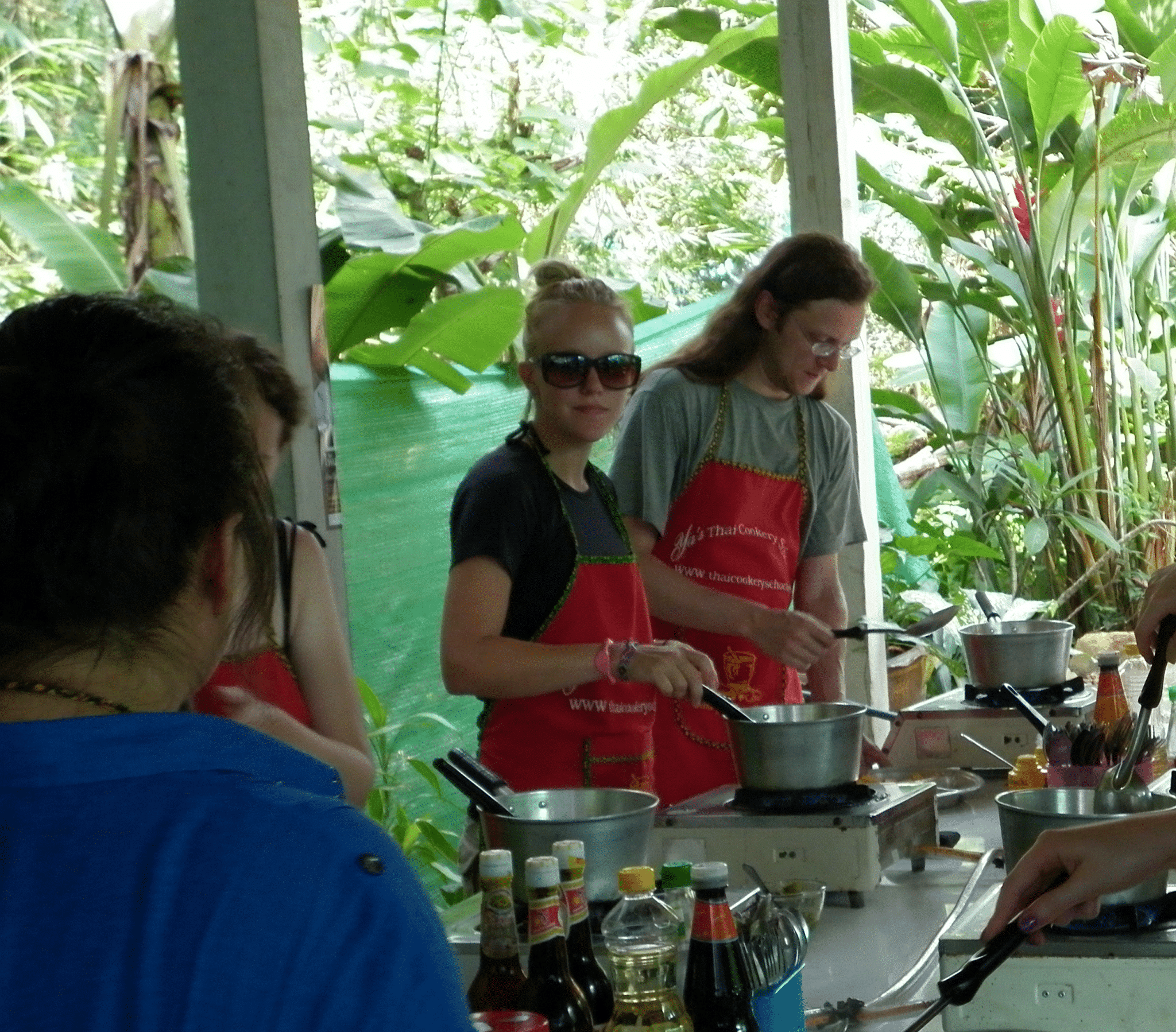 Jessica Randhawa wearing a red chef's apron while cooking at Ya Cookery School in Krabi, Thailand, surrounded by classmates, stovetop burners, pots, and tropical greenery in the outdoor Thai kitchen.
