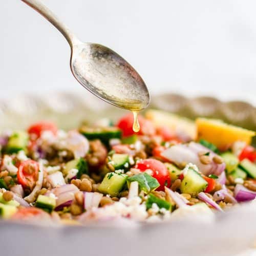 Silver spoon drizzling homemade lemon-Dijon dressing over a salad bowl filled with lentil salad made with with cooked green lentils, fresh parsley, cucumber, feta cheese, red onion, cherry tomatoes.