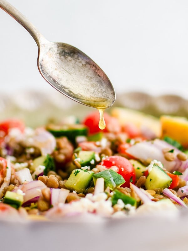 Silver spoon drizzling homemade lemon-Dijon dressing over a salad bowl filled with lentil salad made with with cooked green lentils, fresh parsley, cucumber, feta cheese, red onion, cherry tomatoes.