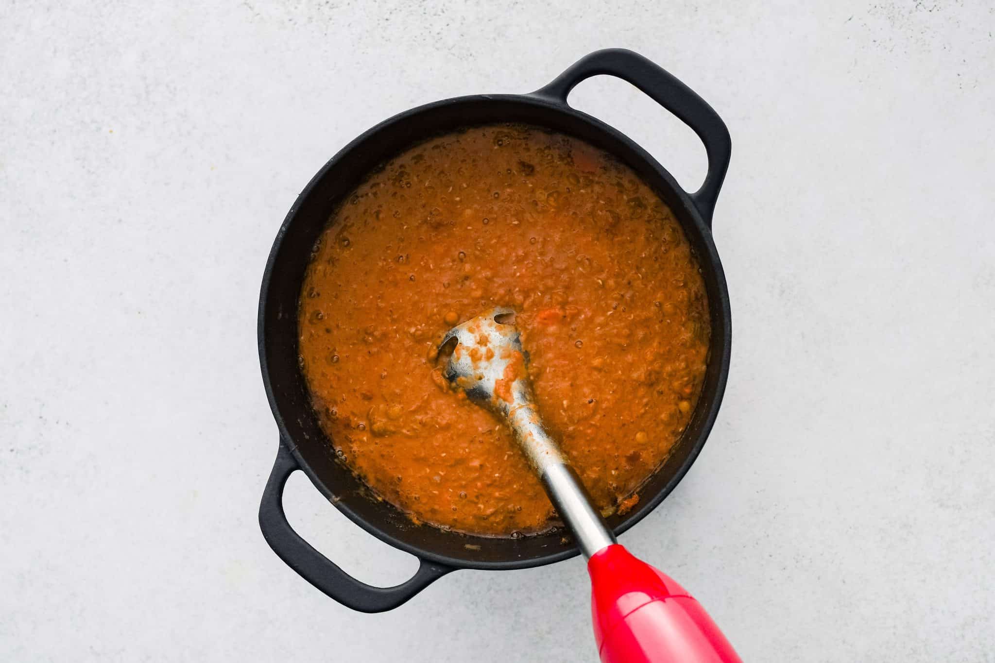 Immersion blender pureeing a portion of lentil soup cooking in a large cast iron pot.