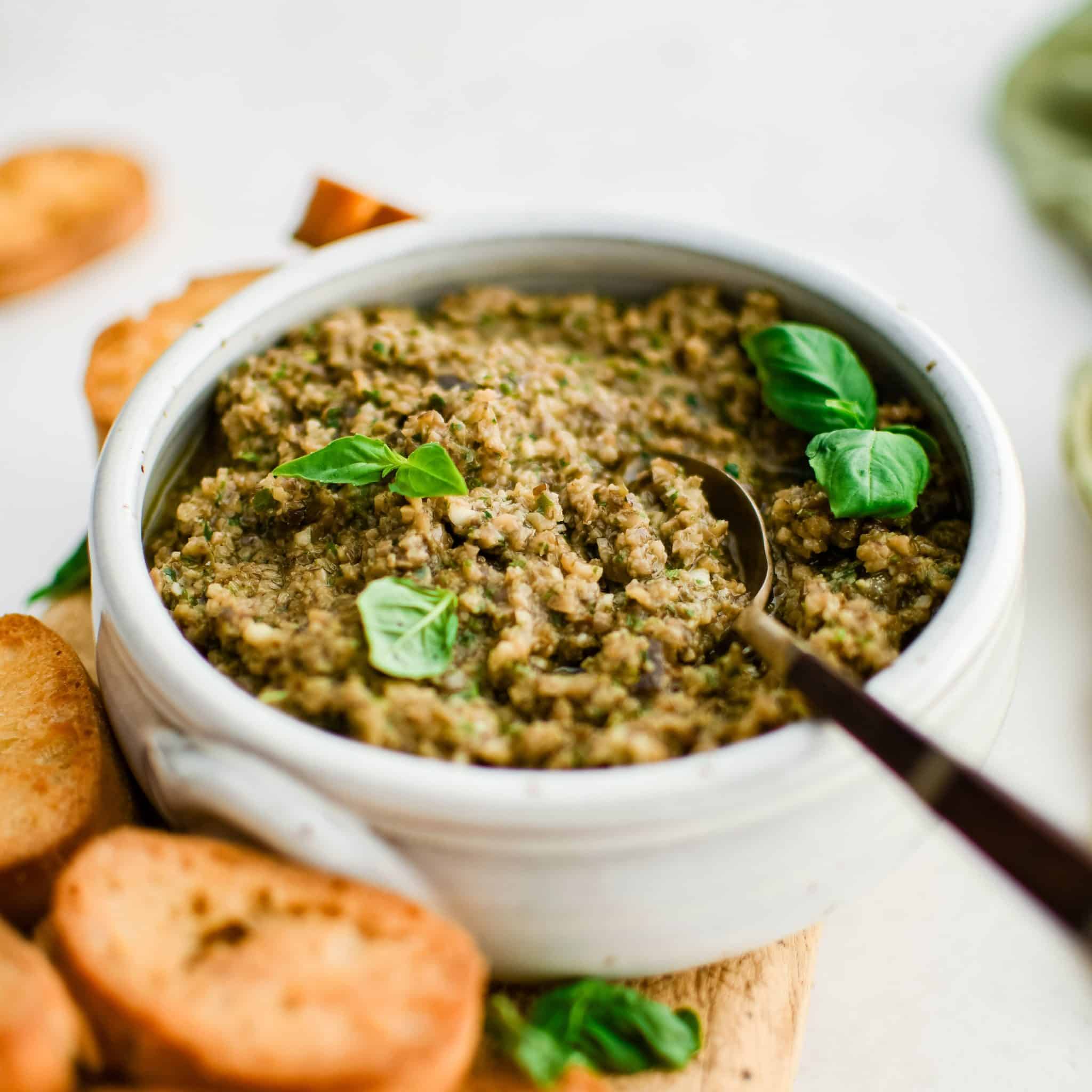 Homemade olive tapenade recipe made with pitted olives, basil, anchovies, parsley, garlic cloves, olive oil. .and capers in a white serving bowl garnished with fresh basil leaves and served with crispy crostini.