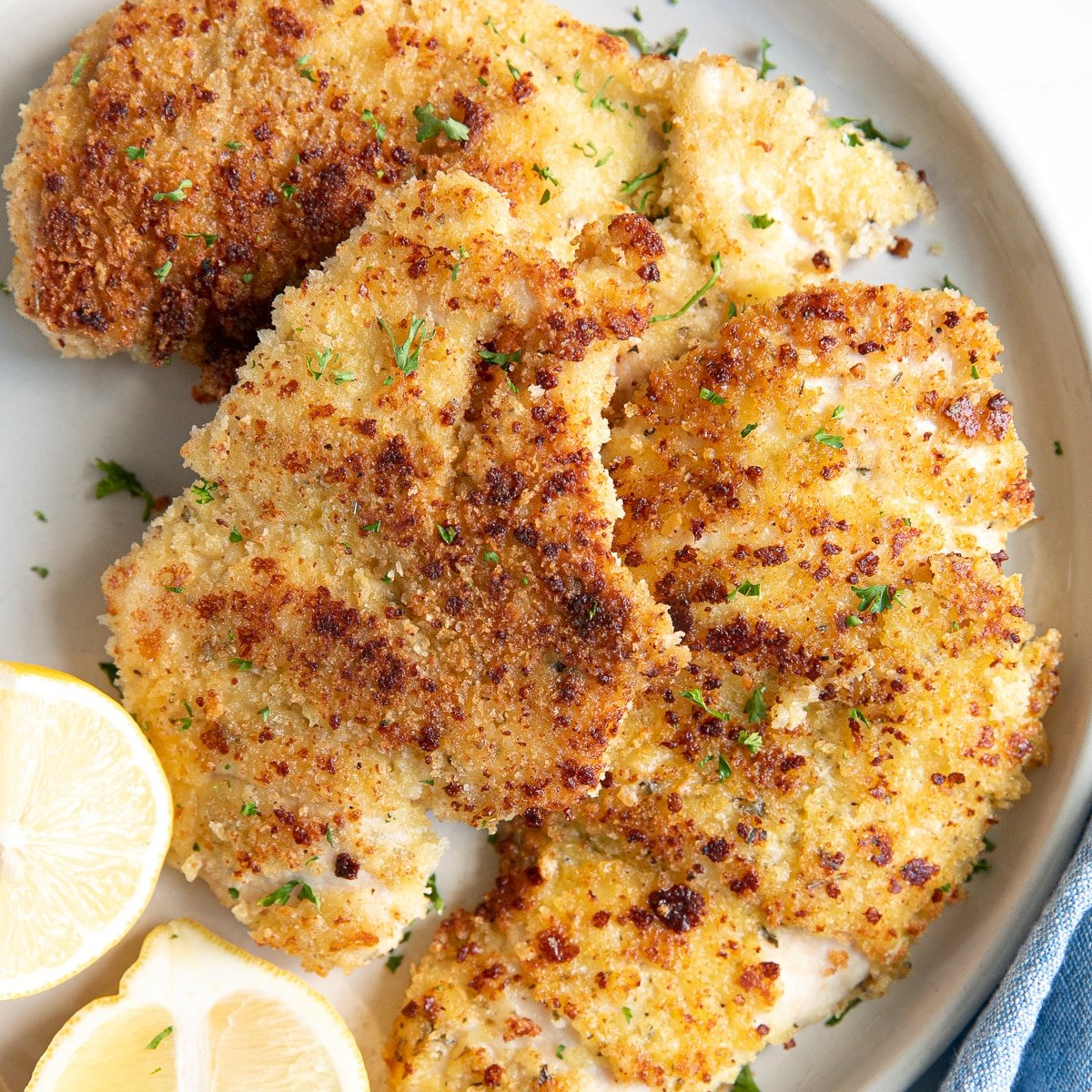 Two thinly sliced parmesan crusted chicken breast pieces on a small white serving plate garnished with chopped parsley and fresh lemon.