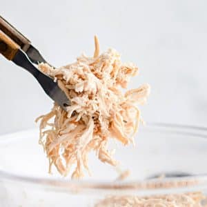 Large forkful of shredded cooked chicken breast hovering above a glass dish filled with additional shredded chicken breast.