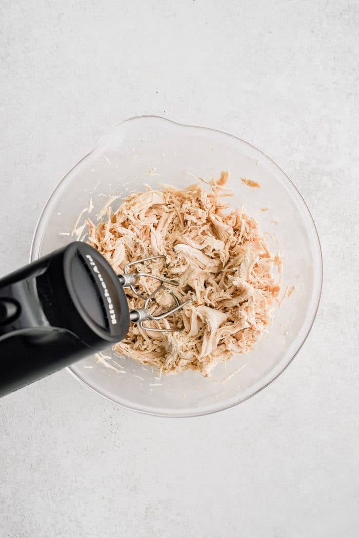 Hand mixer shredding cooked chicken meat in a glass bowl.