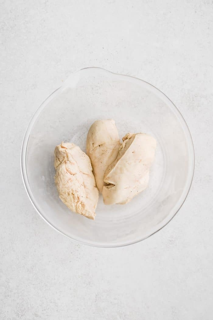 Three large poached chicken breasts in a large glass bowl.