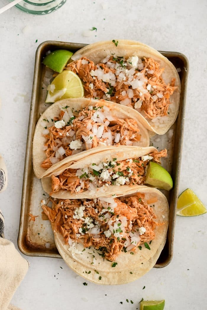 Small baking sheet with four shredded chicken tacos topped with minced white onion and cilantro and served with fresh lime wedges.