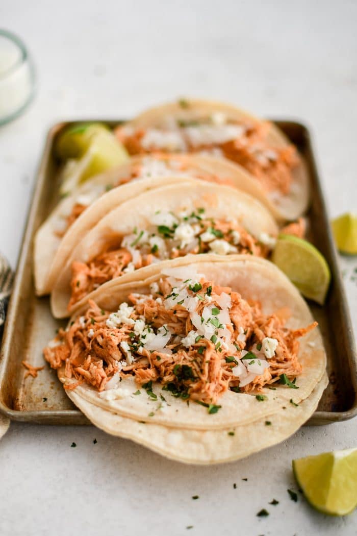 Small baking sheet with four shredded chicken tacos topped with minced white onion and cilantro and served with fresh lime wedges.