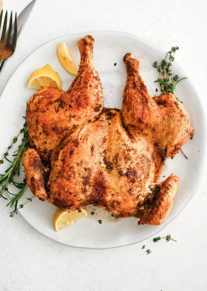 Juicy and golden roasted spatchcock chicken on a white serving platter with lemon wedges and fresh herbs.