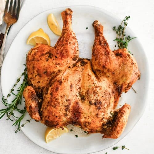 Juicy and golden roasted spatchcock chicken on a white serving platter with lemon wedges and fresh herbs.