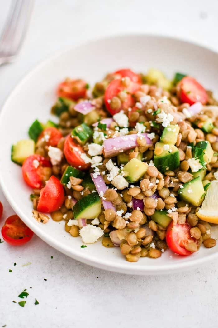 Close-up photo of lentil salad in a shallow white bowl with brown lentils, diced dark green cucumber, halved red cherry tomatoes, chopped parsley, sliced purple-red onion, crumbled white feta cheese, and a lemon wedge on the side.