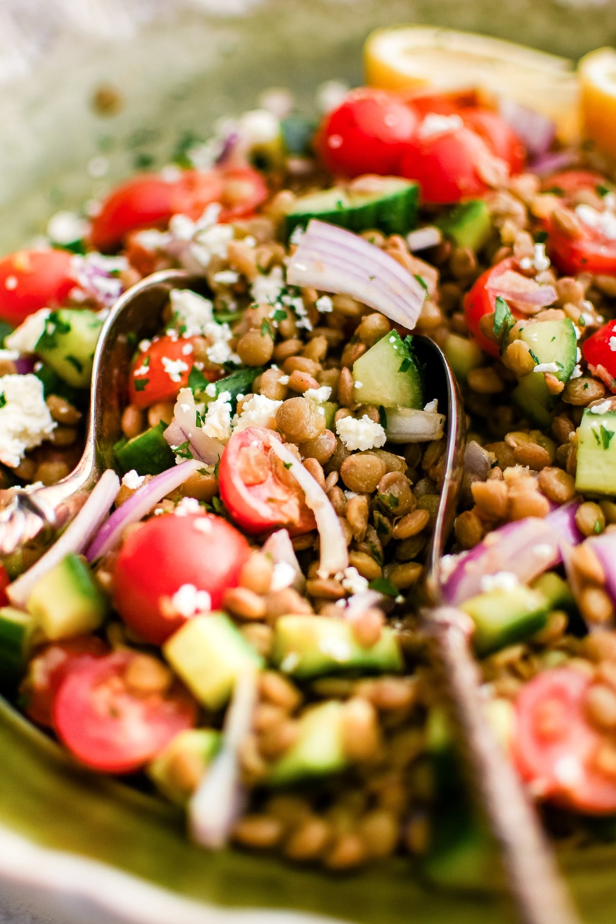 Close-up photo of lentil salad in a light green bowl with a silver serving spoon, showing brown lentils, diced cucumber, halved cherry tomatoes, sliced red onion, chopped parsley, and crumbled white feta cheese. Lemon wedges are blurred in the background.