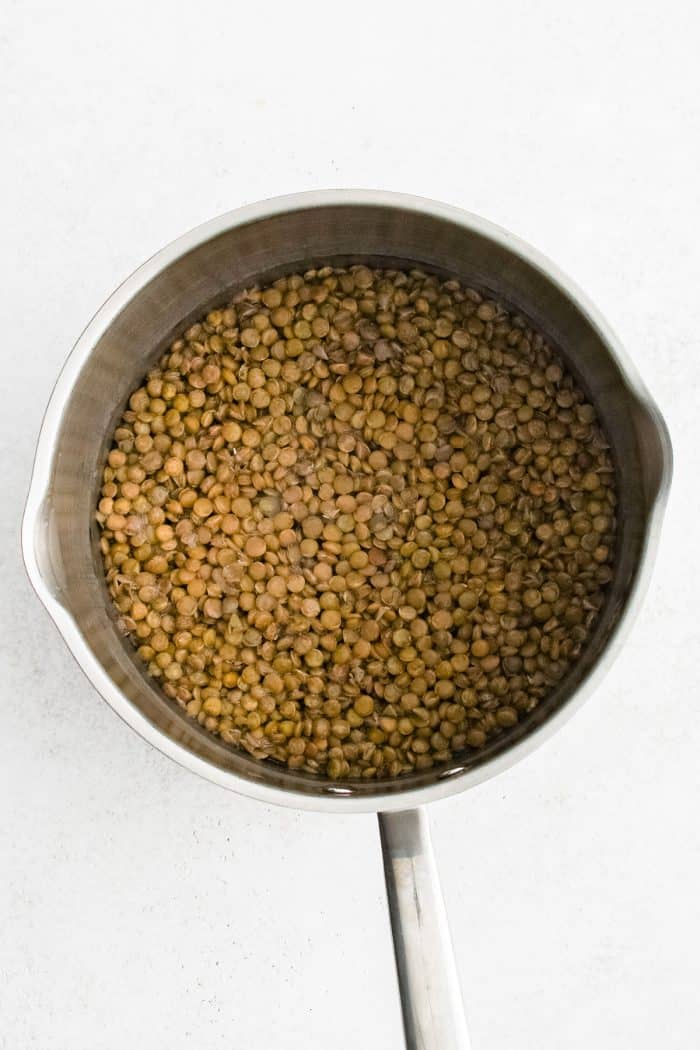 Overhead photo of green-brown lentils simmering in water in a stainless steel saucepan with a long silver handle on a light white background.