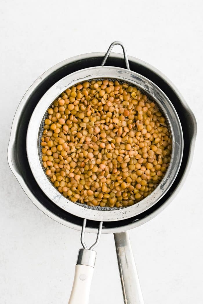 Overhead photo of cooked green-brown lentils drained in a fine mesh strainer set over a stainless steel saucepan with a white-handled strainer on a light white background.
