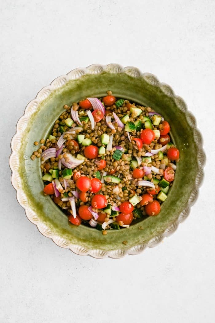 Overhead photo of mixed lentil salad in a scalloped light green serving bowl with cooked lentils, halved cherry tomatoes, diced cucumber, and sliced red onion on a light white background.
